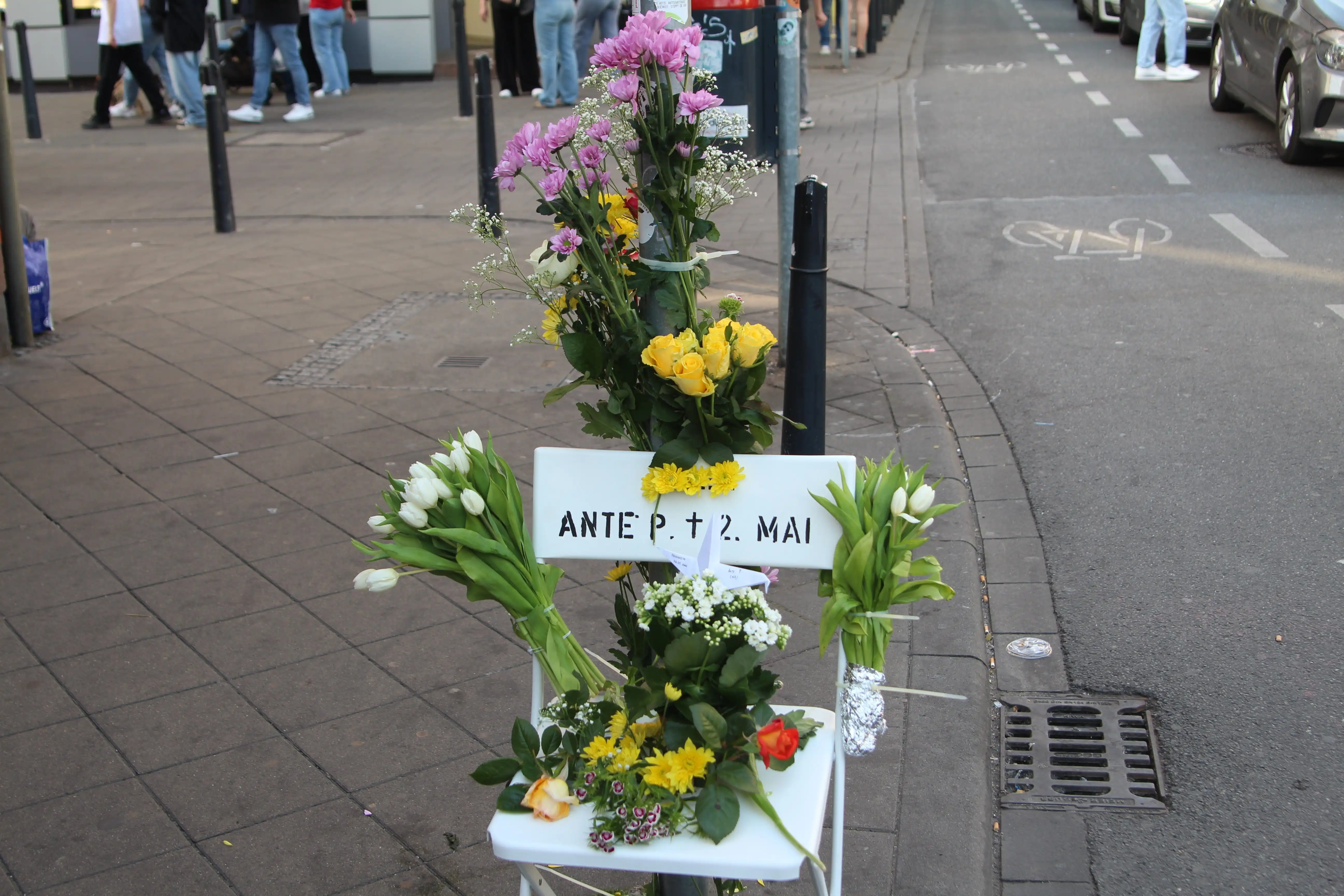 Ein weißer Stuhl, quer über die Rückenlehne aufgemalt in Blockbuchstaben und mit schwarzer Farbe „Ante P. † 2 Mai“.  Auf dem Sitz liegen viele Blumen, auch eine Topfblume steht drauf. Zwischen den Blumen steckt ein gefalteter Papierkranich aus hellem Papier. Unzählige Blumen befinden sich an den Stuhllehnen und dem Pfosten eines Straßenschildes (gegenüber dem Schaufenster der Metzgerei), diese sind mit Kabelbindern befestigt.