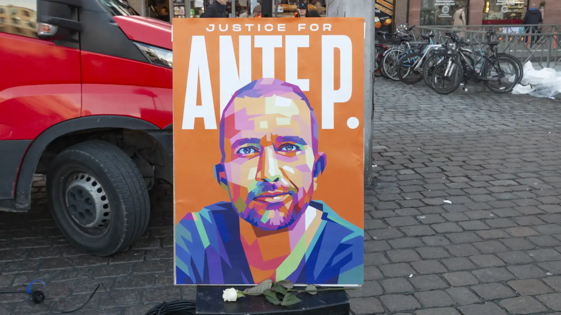 Ein Plakat mit der Aufruf Justice for Ante P. Das Gesicht von Ante P. ist aus bunten 3D-Mosaiken zusammengesetzt. Schrift ist weiß. Hintergrund ist orange. Hinter dem Plakat ist links eine Motorhaube und Rechts Fahrräder zu sehen.