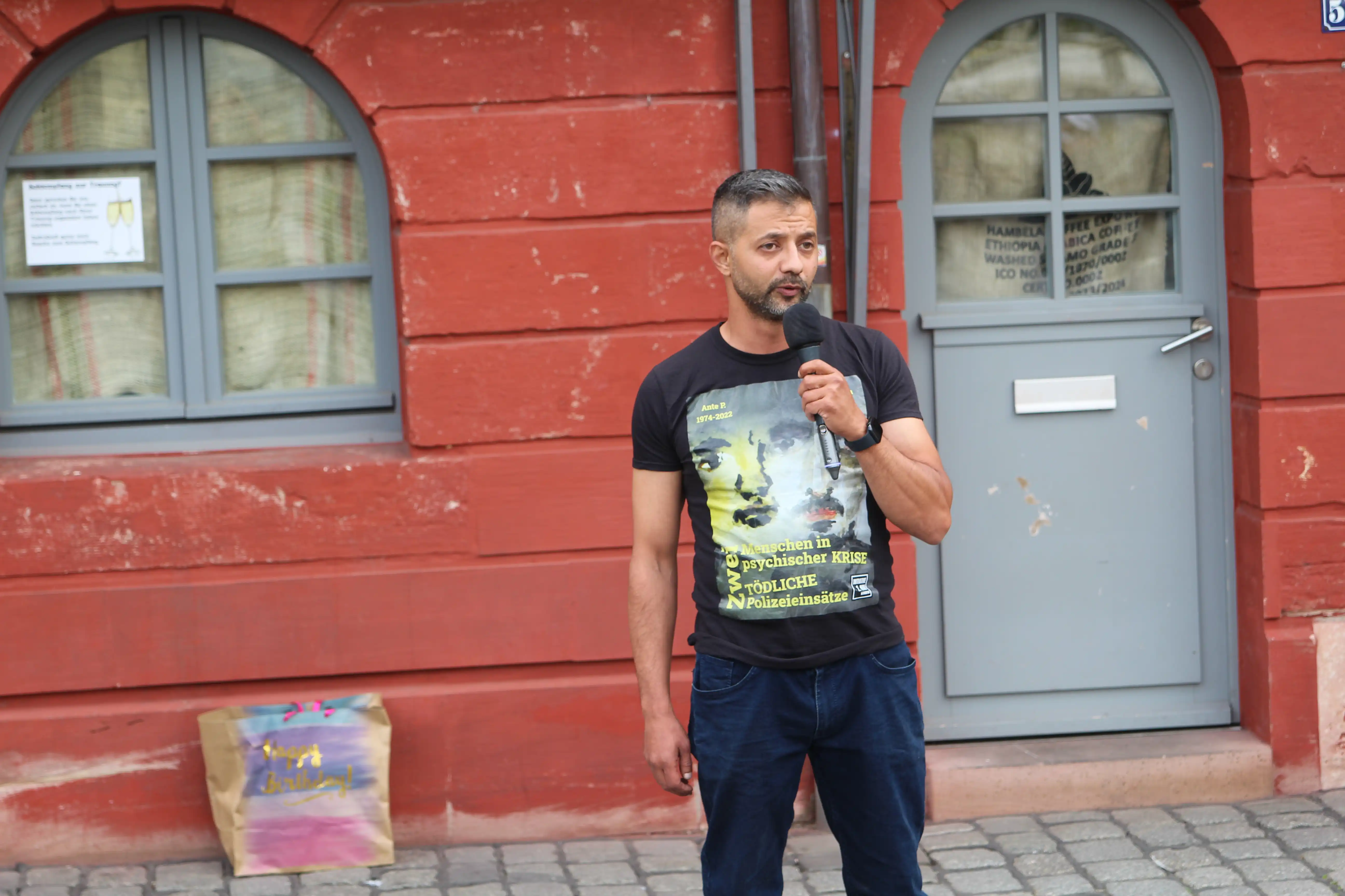 Emrah D. steht auf dem Marktplatz vor einer roten Hauswand. Er hält ein Mikrophon in der Hand und trägt dabei ein schwarzes T-Shirt mit dem Aufdruck der Ausstellung Zwei Menschen in psychischer Krise zwei tödliche Polizeieinsätze mit den Aquarellbildern von Ant und Ertekin. Im Hintergrund ist Links unten im Bild eine Tasche an die Hauswand gelehnt.