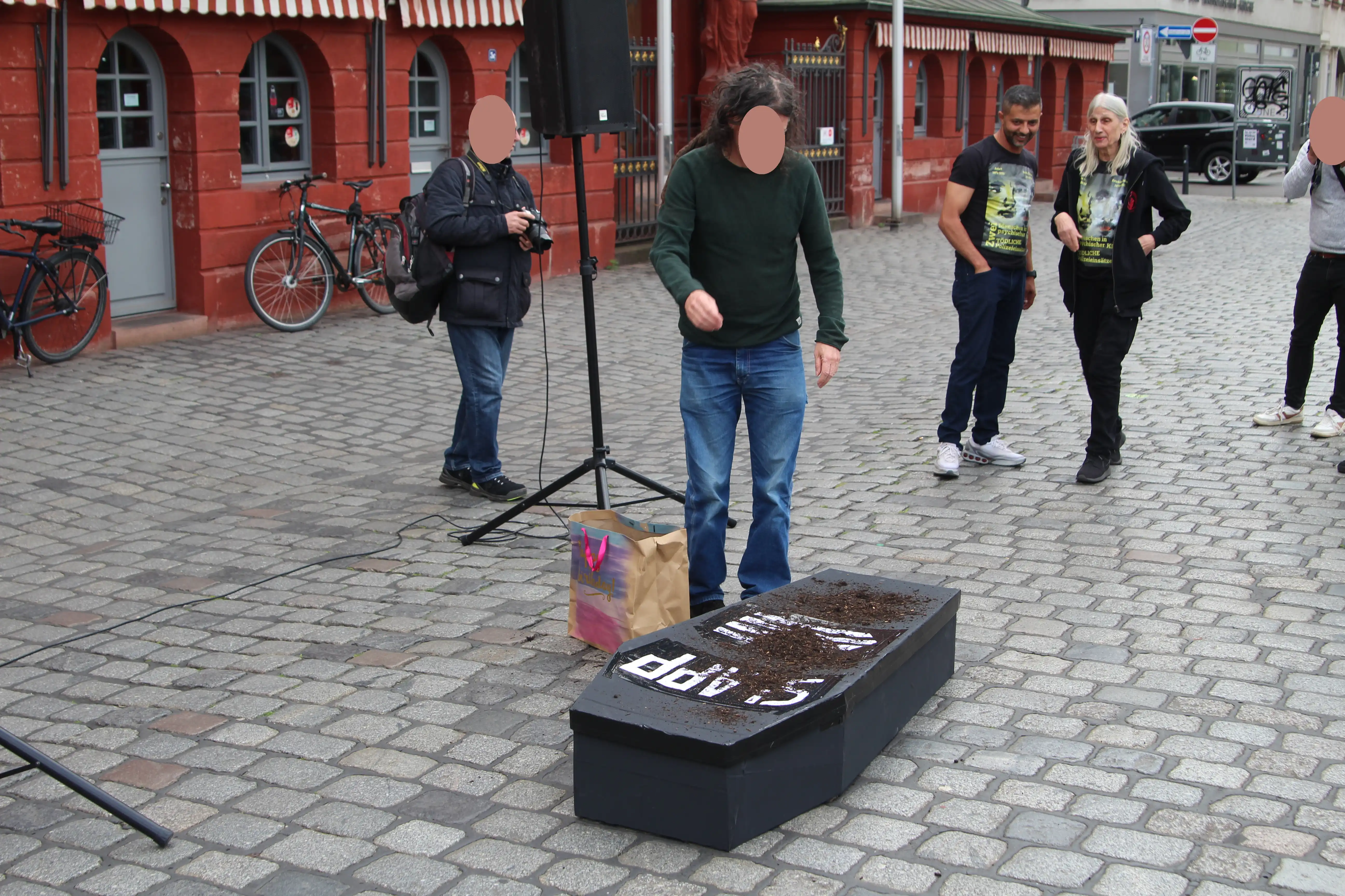 Vor einem mit Erde bedeckten Sarg mit der Aufschrift SLAPP steht eine Person welche gerade Erde auf diesen Sarg wirft. Im Hintergrund stehen noch 4 weitere Personen