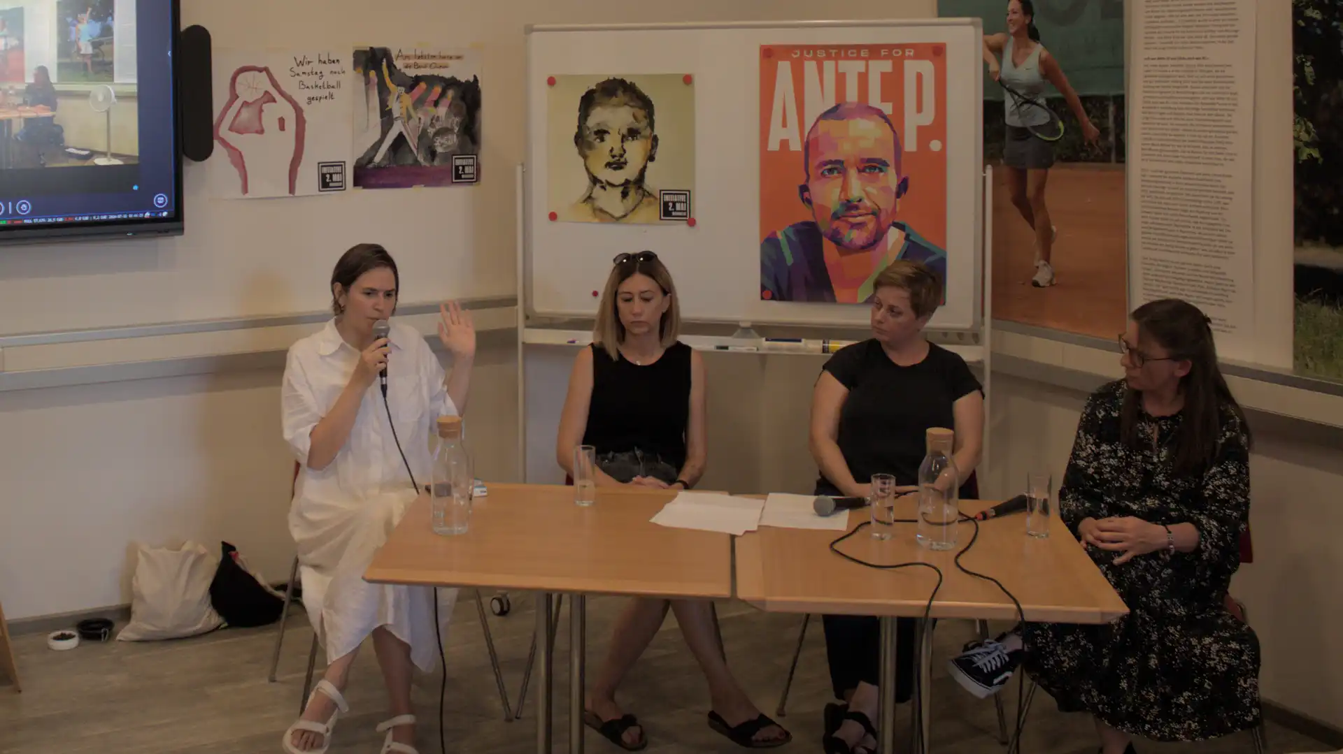 Sevda, in einem weißen Kleid, Antonia und Angelina jeweils in einem einem schwarzen Top und Carina Keppel sitzen an zwei nebeneinandergestellten viereckigen Holztischen. Auf dem Tisch liegen Wassserflaschen und zwei Mikrofone. Sevda hält ein Mikrophon in der HAnd. Im Hintergrund ist links ein kleiner Teil eines Bildschirms zu sehen. An den Wänden und den Aufstellern hängen Poster aus der Ausstellung " Ich will einen Richter" Die Reihe beginnt mit dem Poster Wir haben Samstags noch Basketball geespielt. Daneben hängt das Post Am liebsten hörte er die Band Queen. Auf dem Aufstellwhiteboard hängt ein Aquarellportrait sowie ein Portraitbild mit orangenem Hintergrund und dem Aufdruck Justice for Ante P.