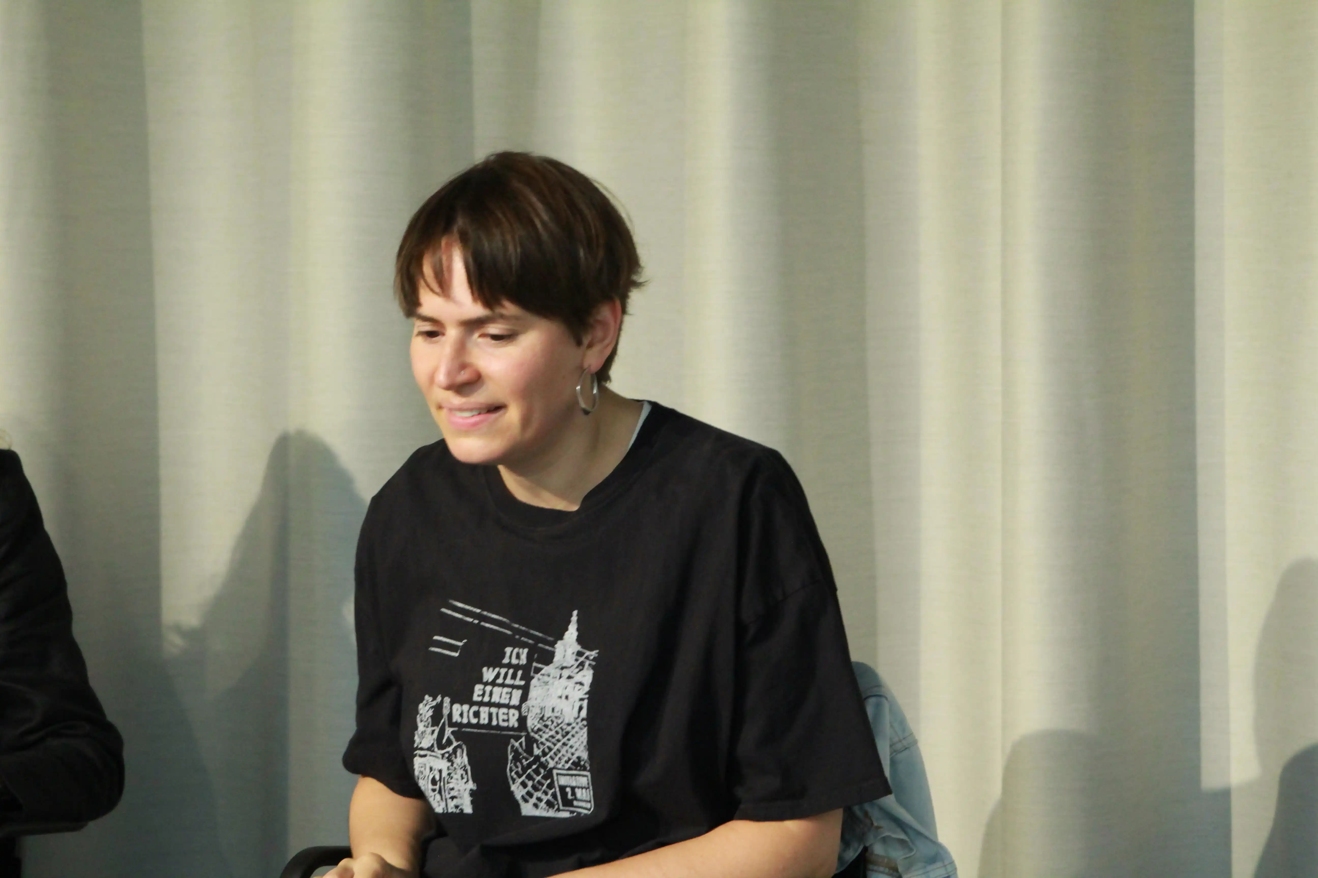 Eine Nahaufnahme von Dr. Sevda Can Arslan. Sie sitzt leicht nach vorne gelehnt vor einem beigen gewellten Vorhang und trägt ein schwarzes T-Shirt mit dem Aufdruck des Ausstellungsbildes „Ich will einen Richter“