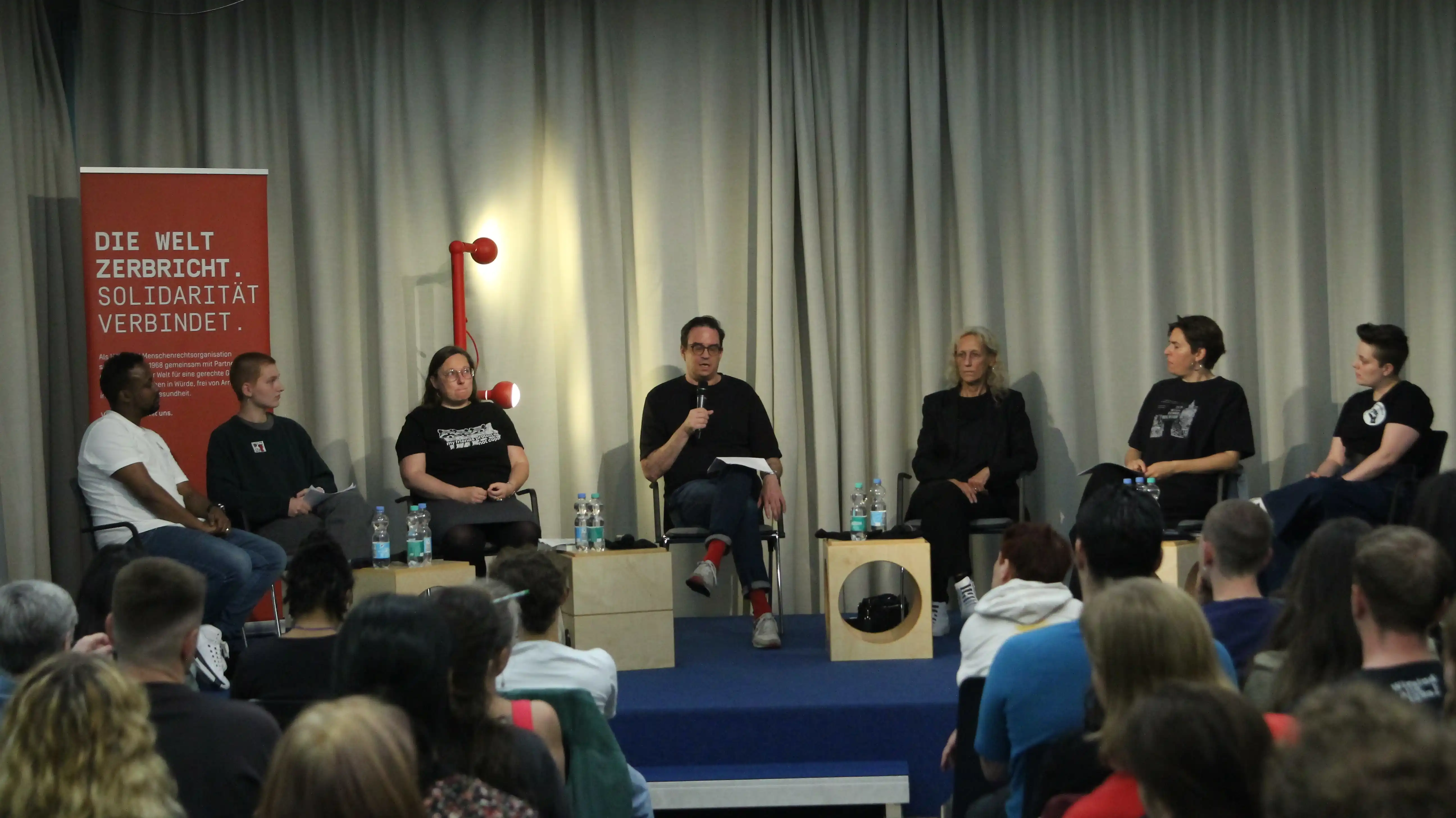die Gäst:innen auf dem Podium, es sprechen von links nach rechts: Kamal und Klee vom Solikreis Biriq; Michelé Winkler vom Grundrechtekomitee; Moderator Daniel Loick; Rechtsanwältin Beate Böhler; Dr. Sevda Can Arslan von der Initiative 2. Mai und Teddy von Justice 4 Mouhamed Dahinter ein beiger Vorhang, beleuchtet von 2 Lampen an dunkelrotem Ständer. Links ein dunkelrotes Roll up, zu erkennen weißer Schriftzug „Die Welt zerbricht, Solidarität verbindet“. Darunter steht noch mehr, aber zu klein zum Erkennen was da noch geschrieben ist. Im Vordergrund: Rücken und Hinterköpfe von zuhörenden Menschen.