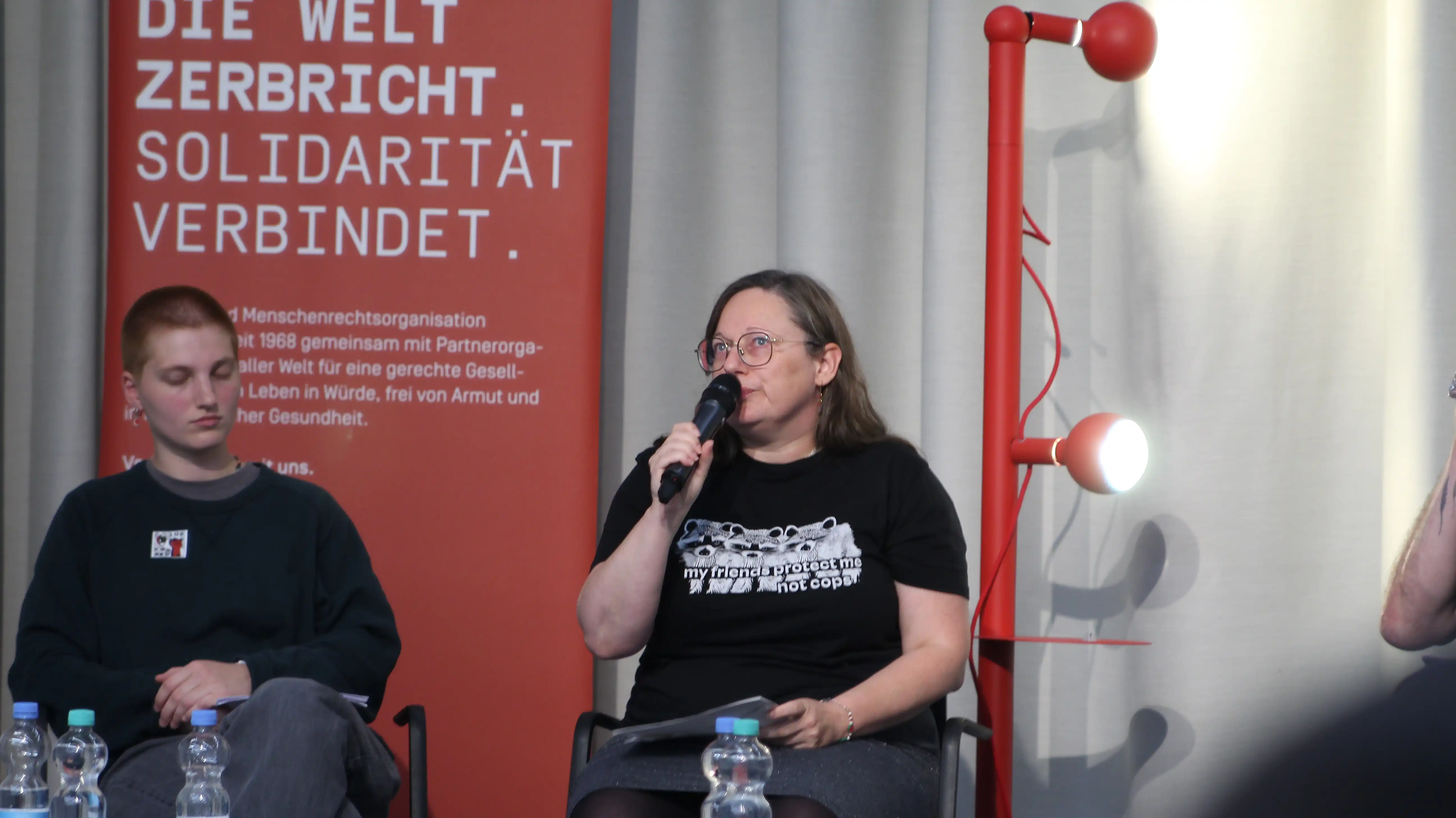 Hier nochmal Klee und Michelé die wieder ins Mikro spricht. Links hinter ihnen ein roter Aufsteller mit der Aufschrift „Die Welt zerbricht, Solidarität verbindet“