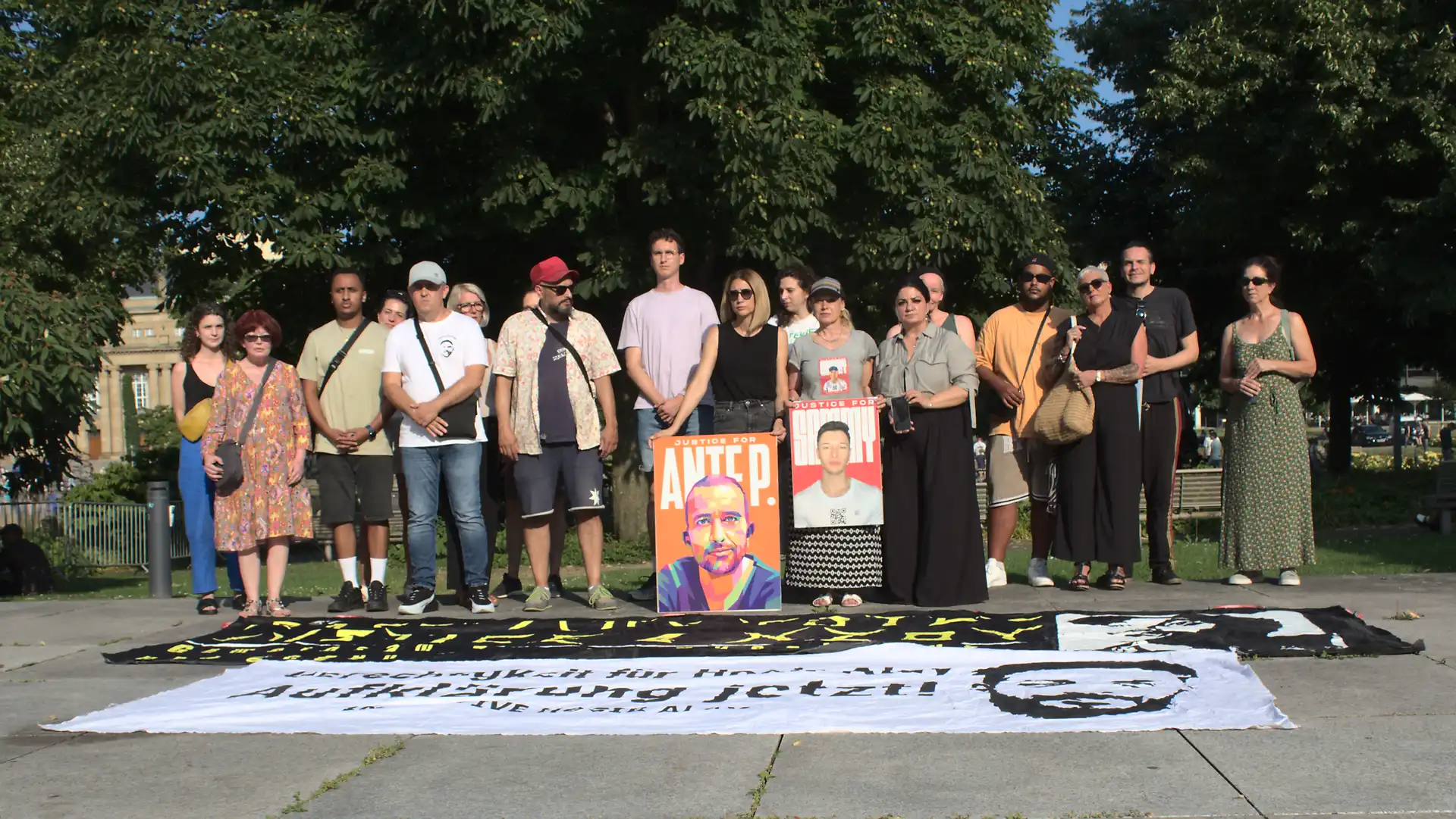 Zusammen mit dem Vater von Hamza Kurtović, den Eltern von Sammy Baker, der Schwester von Ante, den Initiativen Oury Jalloh, Hogir Alay, Nzoy und vielen mehr haben wir uns versammelt. Hinter uns liegt der Landtag von Baden-Württemberg. Vor uns liegen zwei Transparente. Die Gruppe hält Plakate mit den Gesichtern von Ante P. und Sammy Baker. Auf dem ersten Transparen lässt sich der Schriftzug Gerechtigkeit für Hogir Alay Aufklärung jetzt erkennen